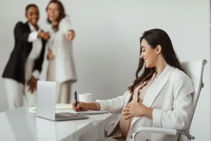 A pregnant woman working at her desk while her coworkers laugh at her.