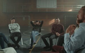 A group of people sit in a wide circle on chairs inside a dimly lit indoor sports gymnasium or basketball court.