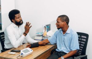 A healthcare professional with a beard, wearing a white button-up shirt, checks a young man's blood pressure using a digital monitor on a wooden desk during a medical consultation.