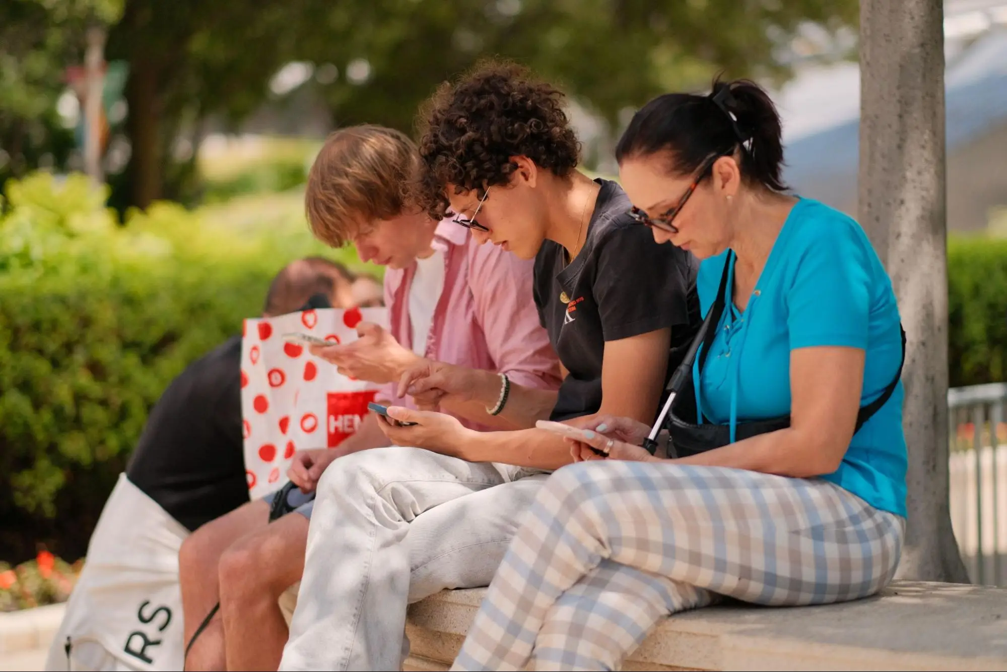 Three people are sitting outdoors on a bench while closely looking at their mobile phones.