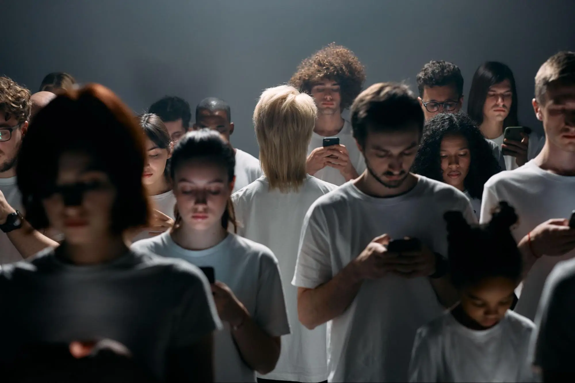A crowded, diverse group of people standing close together in a dimly lit space, with each individual looking down, fully absorbed in using their smartphone.