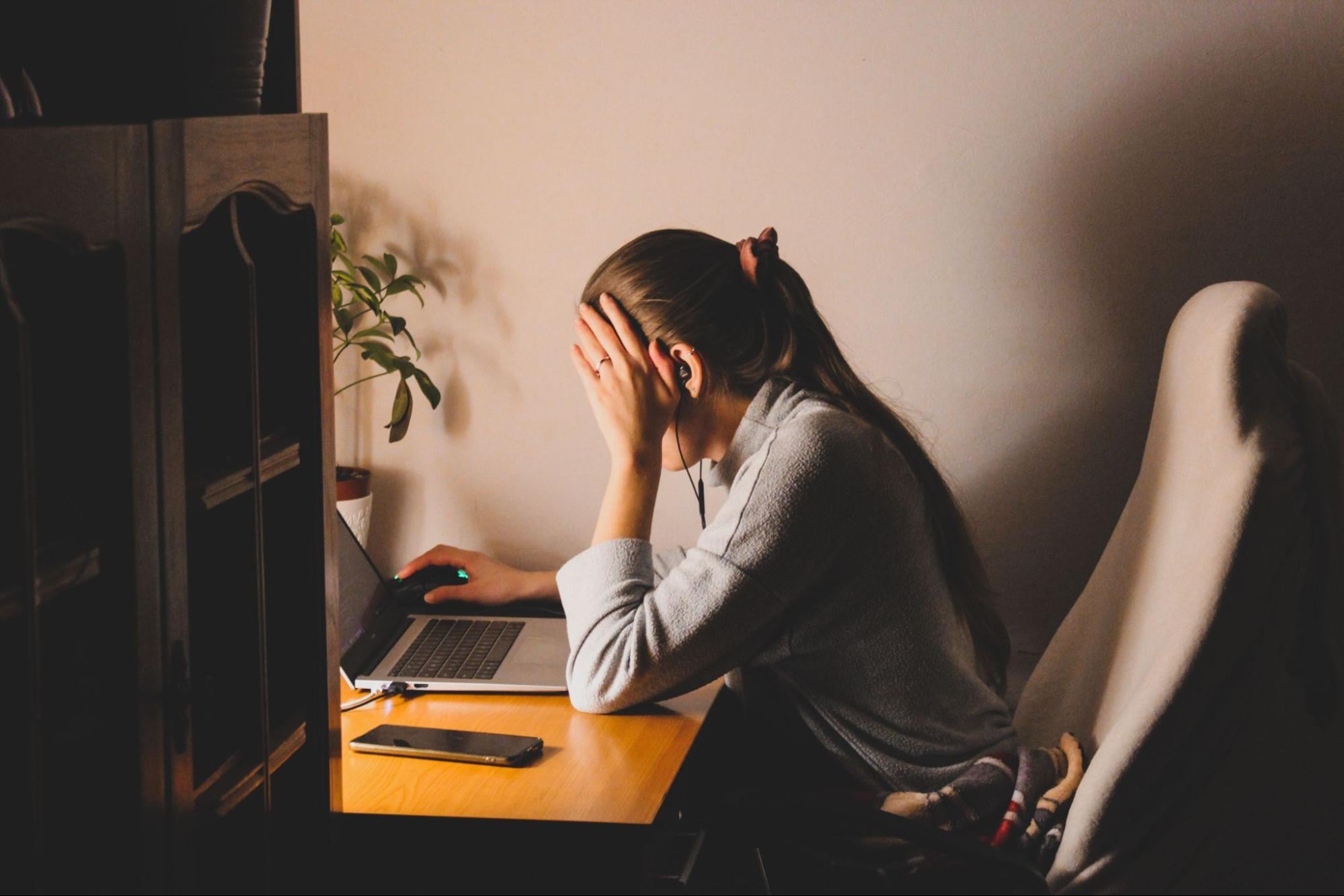 A person wearing earbuds sits at a desk in a dimly lit room, holding their head in one hand while using a laptop, with a phone and plant nearby.