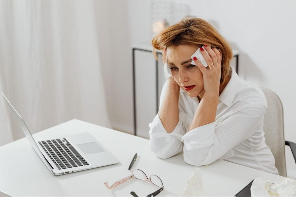 A person with red hair in a white blouse sits at a white desk holding their head in their hands beside an open laptop, glasses, and crumpled tissues.