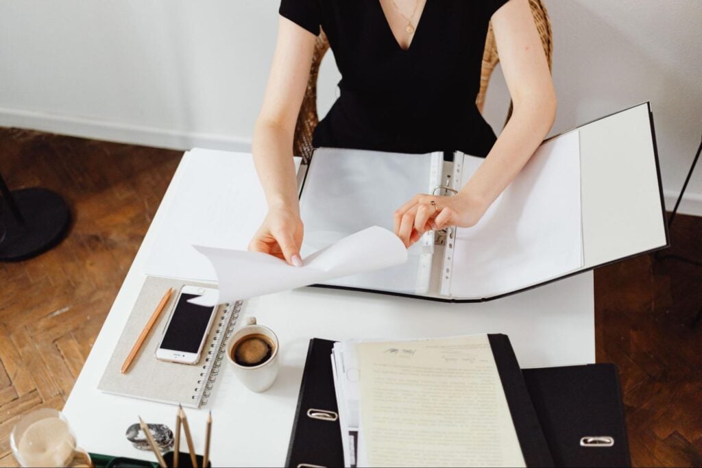 A person in a black top sits at a white desk flipping through pages in an open ring binder, with pencils, a coffee cup, and more binders nearby.